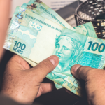 Mãos segurando notas de 100 reais, com cesto ao fundo.