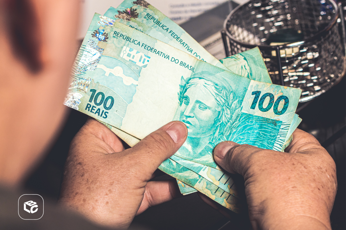 Mãos segurando notas de 100 reais, com cesto ao fundo.
